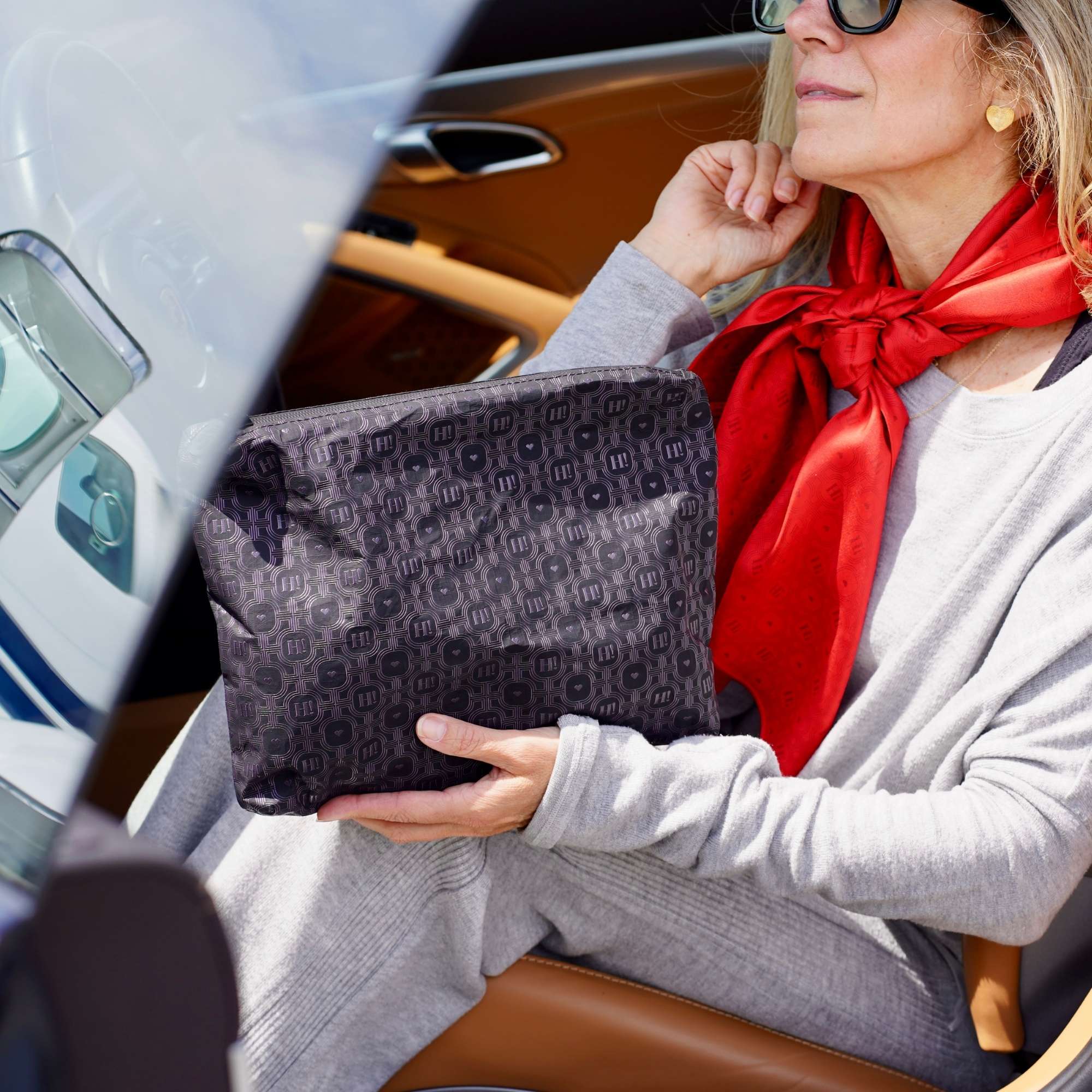 A woman in a gray sweater and red scarf holds a black signature patterned pouch inside a car with tan leather seats.