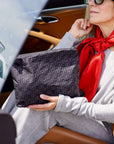 A woman in a gray sweater and red scarf holds a black signature patterned pouch inside a car with tan leather seats.
