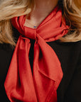 A close-up of a woman wearing a vibrant red H print silk scarf tied around her neck, styled with a black textured top and a small blue heart necklace.