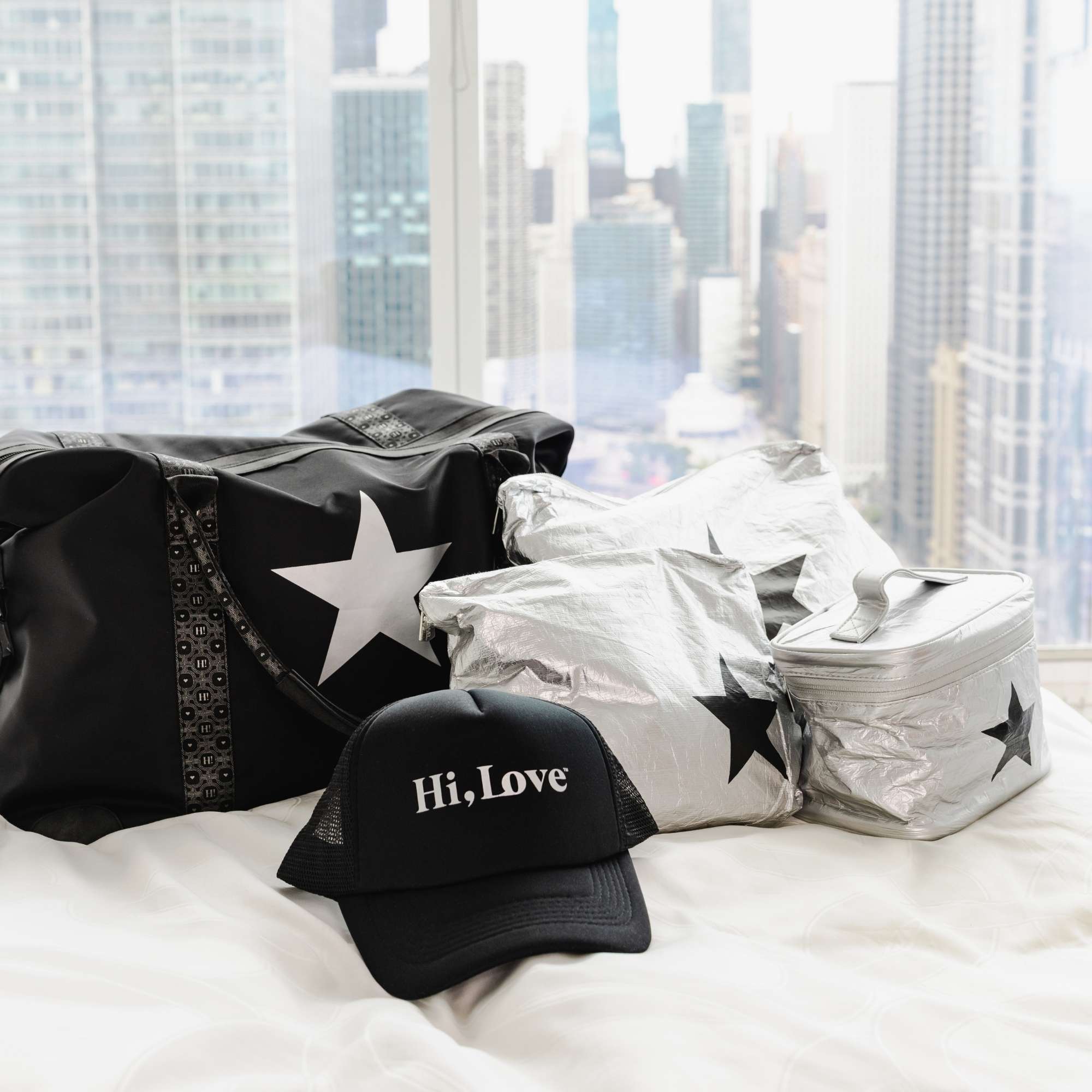 Black Signature Weekender with a white star beside silver Jumbo and Medium Pouches and a silver Cosmetic Carrier Lunch Box with black stars and  Hi, Love Trucker Hat lying on a bed with a city scape background.