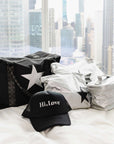 Black Signature Weekender with a white star beside silver Jumbo and Medium Pouches and a silver Cosmetic Carrier Lunch Box with black stars and  Hi, Love Trucker Hat lying on a bed with a city scape background.