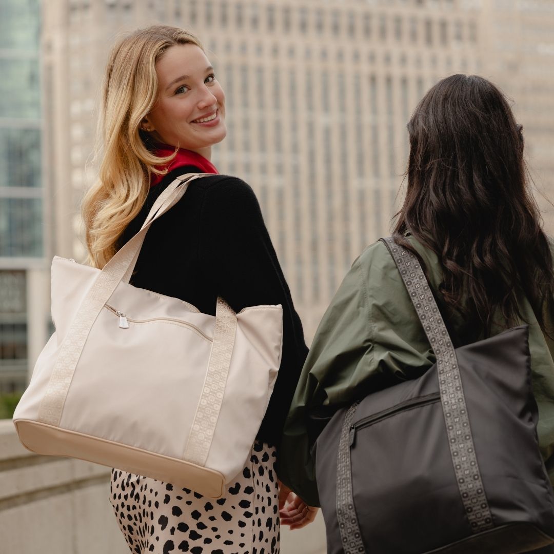 Two women walking outdoors in an urban setting, each carrying a large tote bag. The woman on the left wears a beige tote bag with beige trim, while the woman on the right wears a black tote bag with a gray patterned strap.