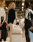 Travelers at a train station with the Signature beige travel tote and matching medium pouch on a carry-on suitcase.