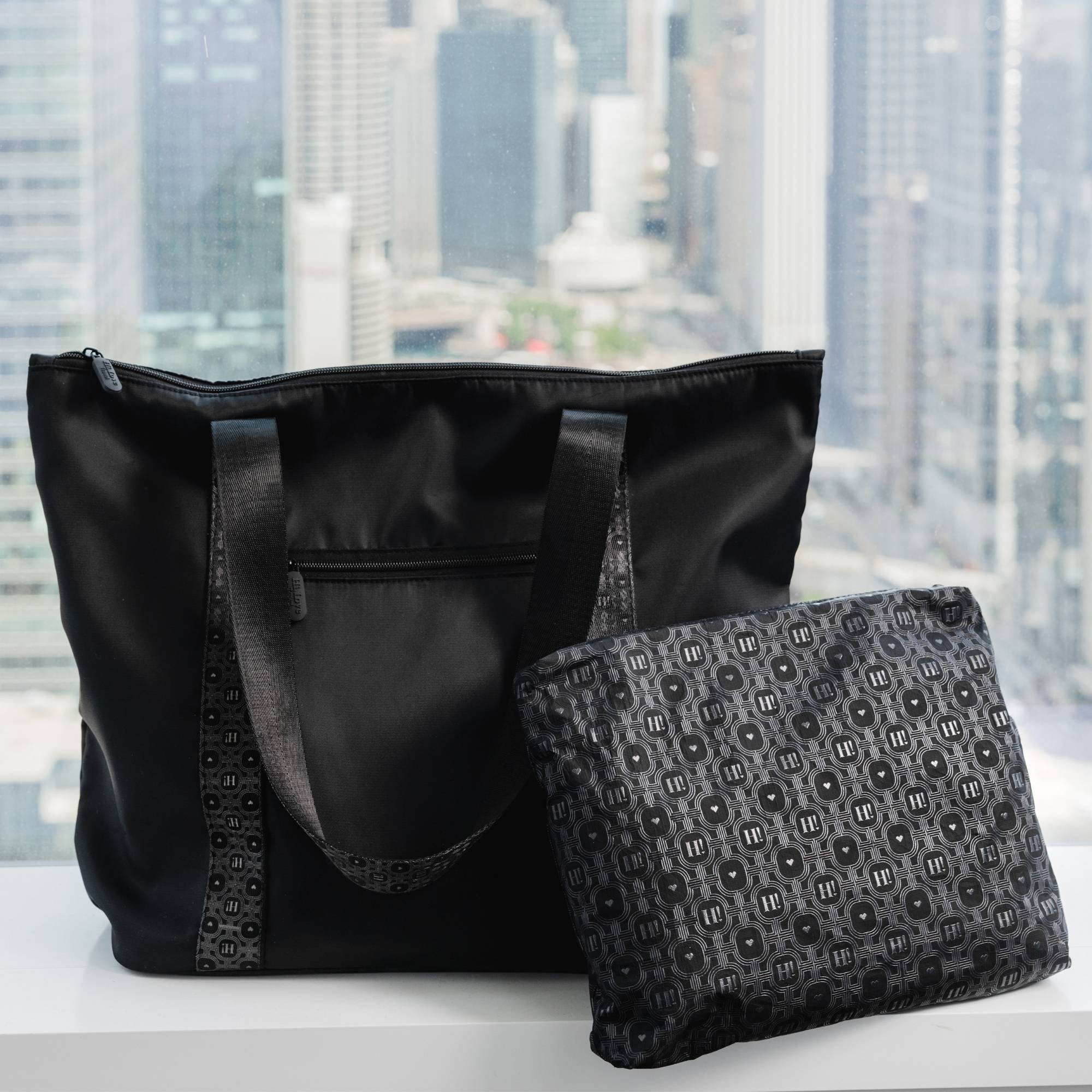 Black signature tote and matching zip pouch set on a bed with a city skyline view behind.