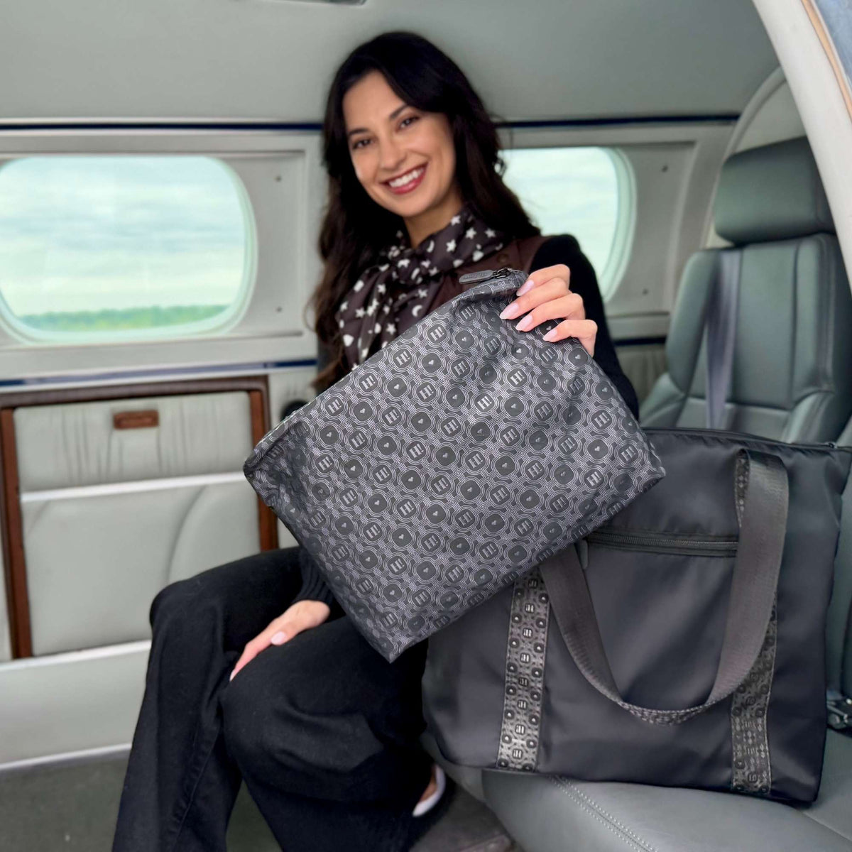 A woman with a scarf sits inside a plane, holding a black signature tote with patterned fabric lining and handles with a matching medium pouch