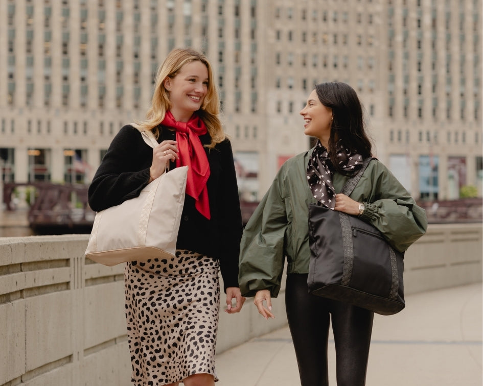 Two women are walking and smiling outdoors on a city bridge, each carrying a large tote bag. The woman on the left carries a beige tote bag, while the woman on the right carries black zippered tote bag.