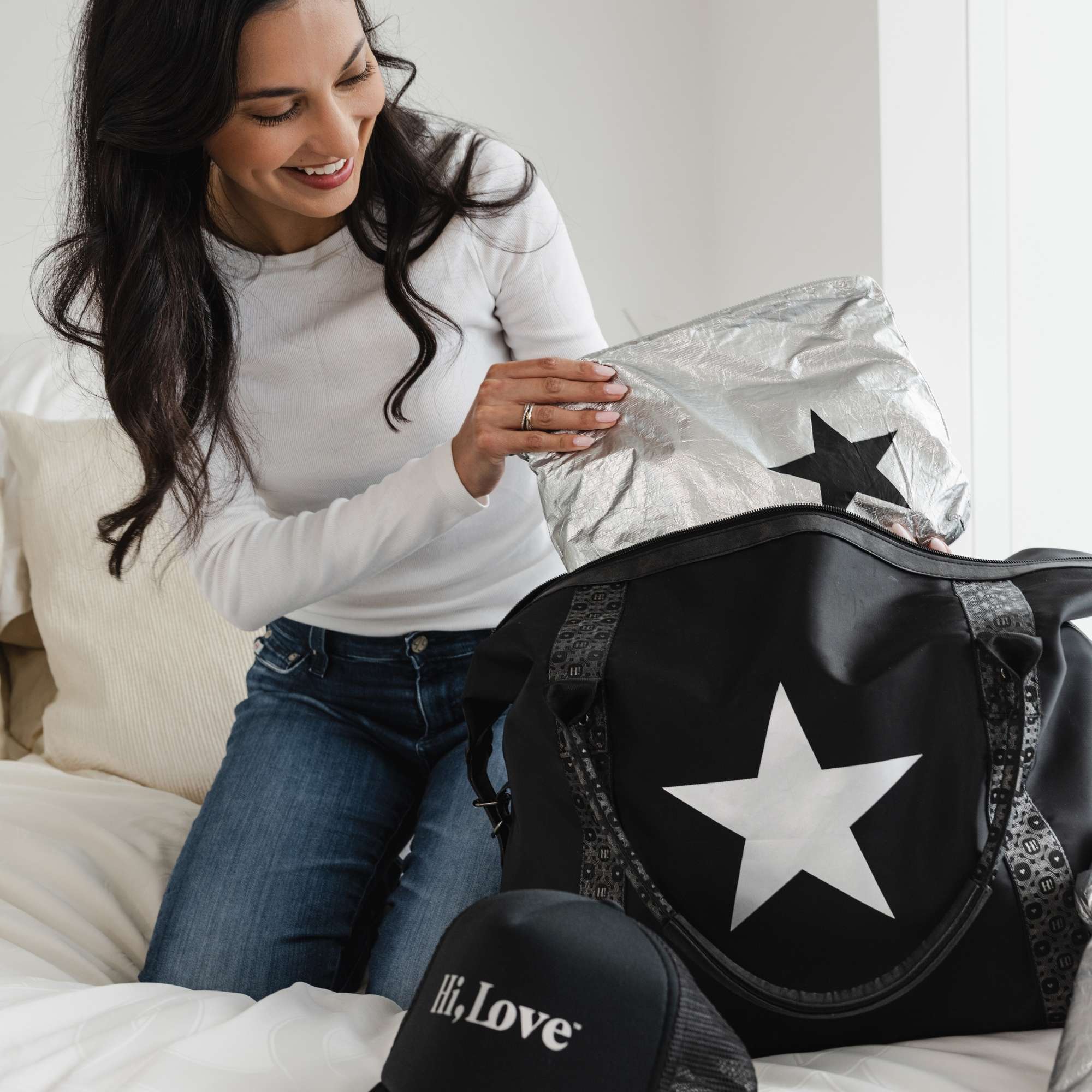 Woman smiling while packing a black Signature Weekender with a silver star and holding a silver medium Pouch with a black star.