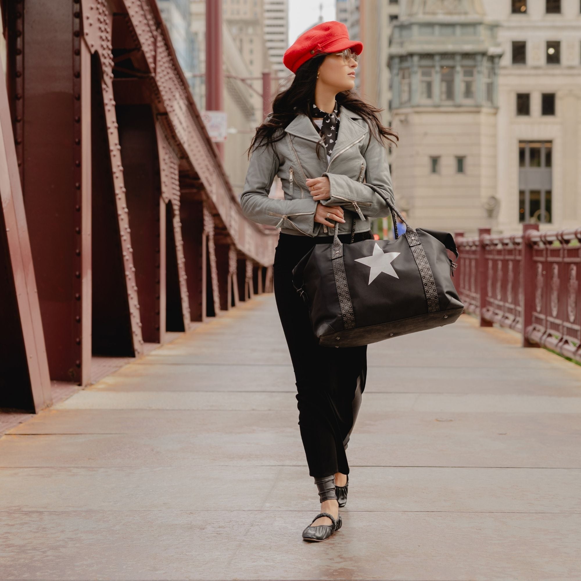 Woman walking with the Signature Weekender Star travel tote bag on a city bridge wearing long black skirt, gray jacket and red hat.