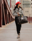 Woman walking with the Signature Weekender Star travel tote bag on a city bridge wearing long black skirt, gray jacket and red hat.