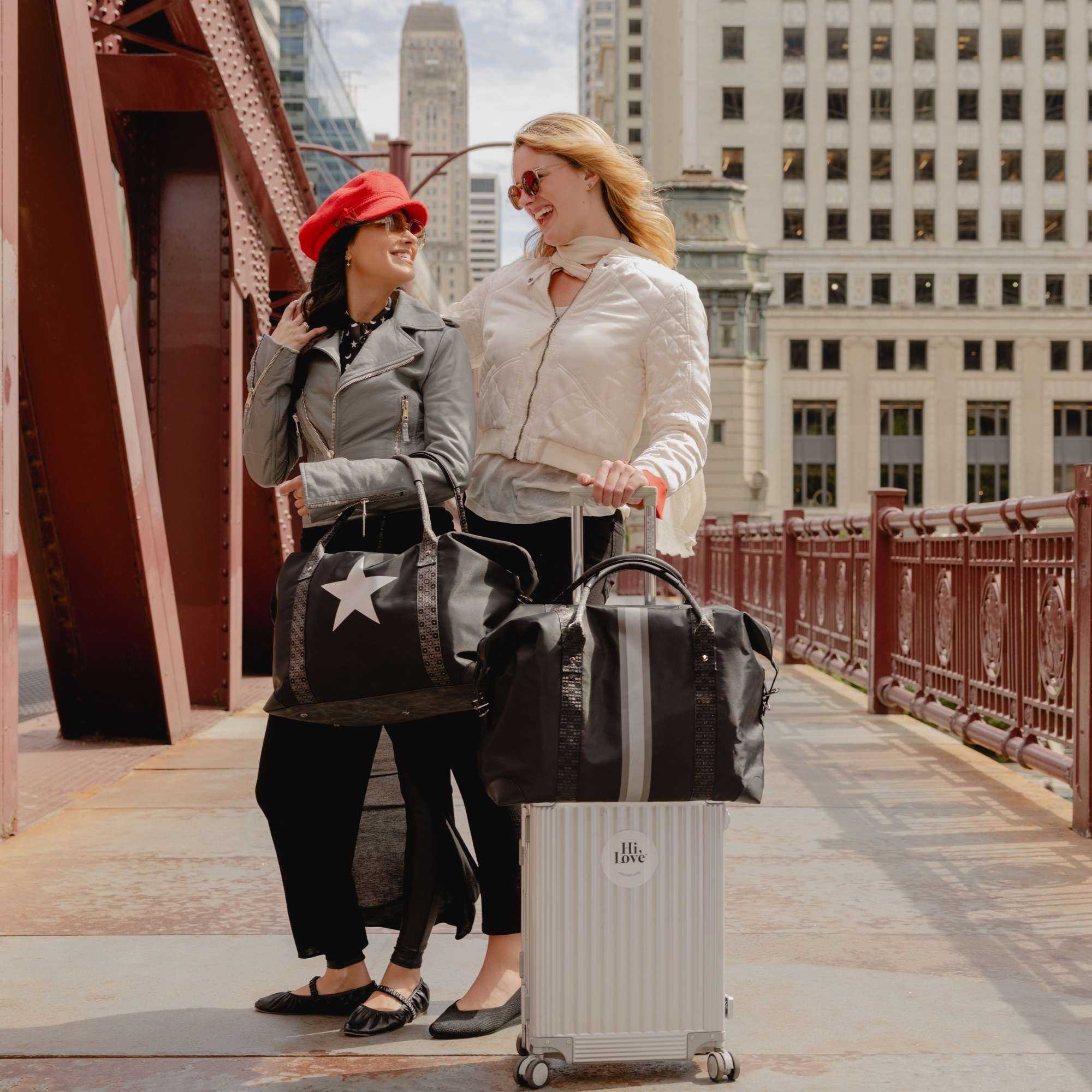 Two women traveling in the city with black weekender bags featuring a pewter and silver stripe accompanied by a coordinating black silver star weekender.