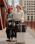 Two women traveling in the city with black weekender bags featuring a pewter and silver stripe accompanied by a coordinating black silver star weekender.
