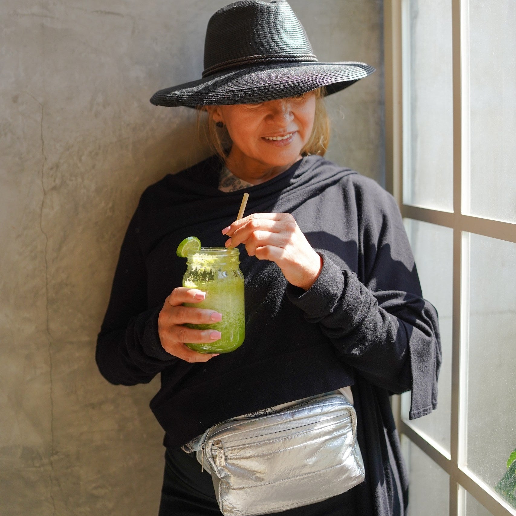 Person wearing a black hat and black outfit with silver fanny pack, holding a green drink with a straw, standing against a neutral wall.