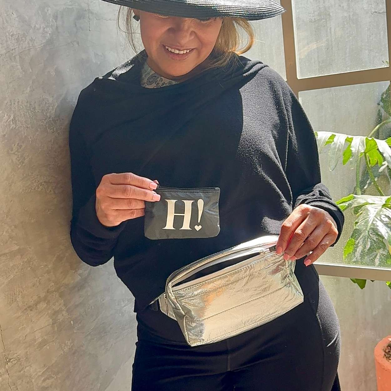 Person holding a card with 'H!' on it, wearing a black outfit, silver fanny pack and a hat, standing indoors.