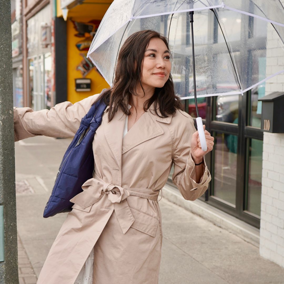 Woman standing on sidewalk holding clear umbrella and navy blue love me knot tote bag wearing a tan trench coat