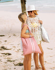 Mother and daughter standing on the beach looking toward each other. One wearing a pink tote bag and the other wearing a silver tote bag.