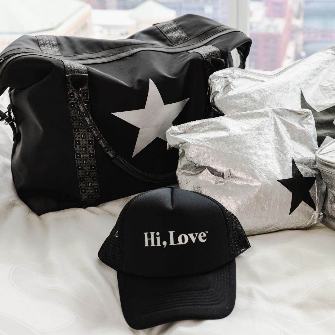 A black silver star duffel bag, a matching black trucker cap with 'Hi, Love' in white, and several silver pouches in star-patterned, arranged on a white bed.