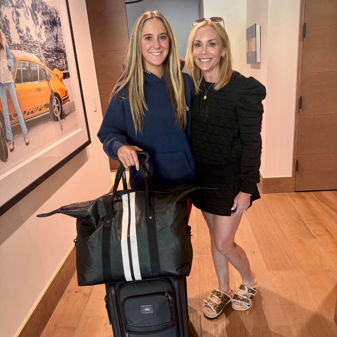 Two women standing in a room with a painting of a yellow taxi on the wall, with a suitcase and a black weekender bag.