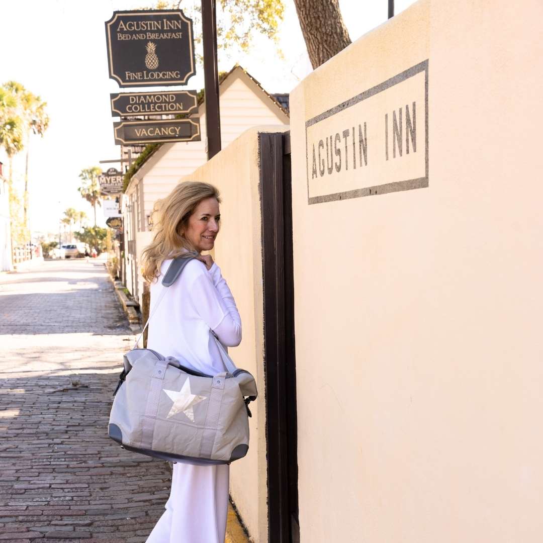 A woman holding a gray weekender bag with a star design in front of the Agustin Inn.