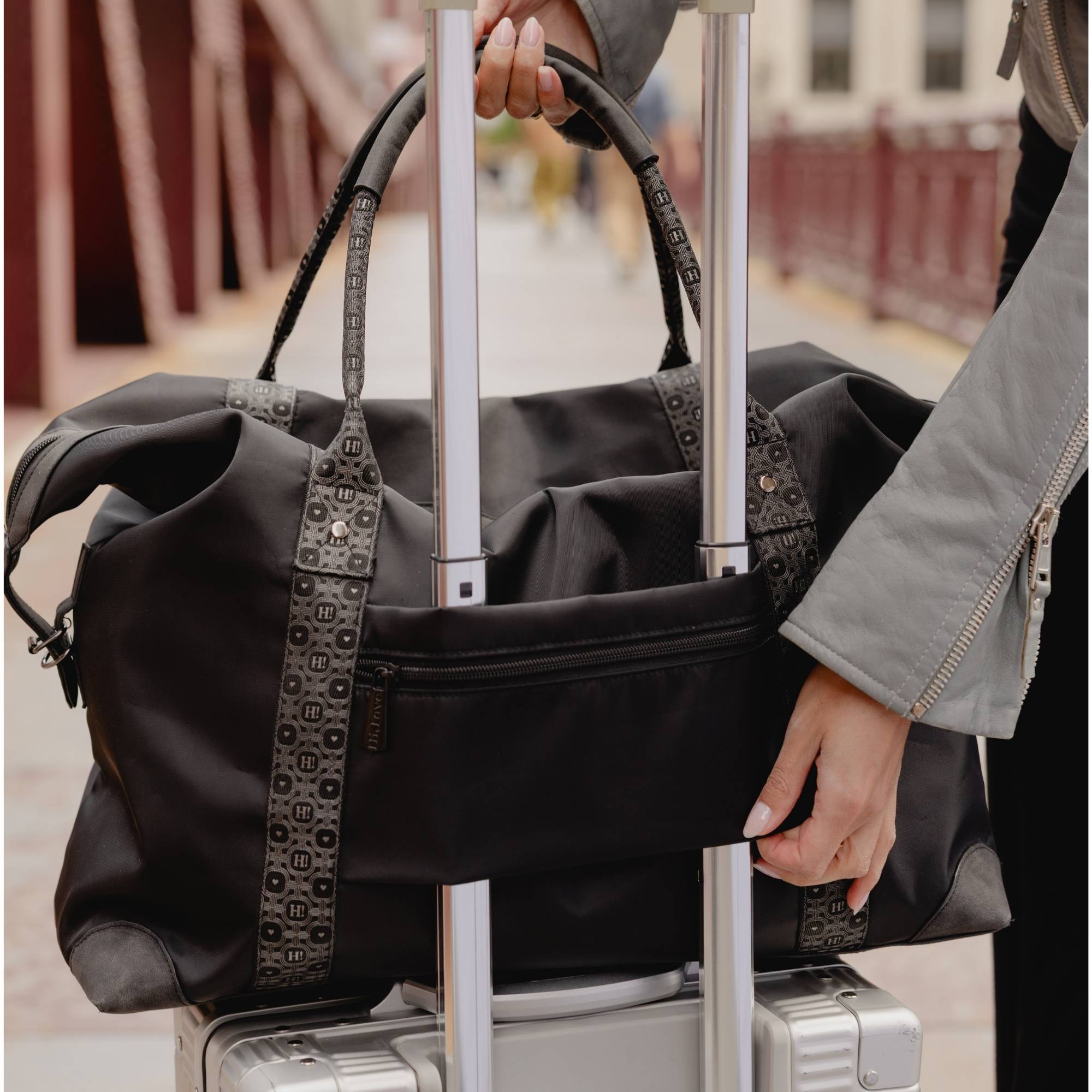 Black weekender duffle bag with silver star and signature straps shown on a suitcase trolley sleeve.