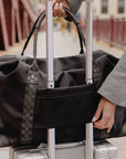 Black weekender duffle bag with silver star and signature straps shown on a suitcase trolley sleeve.