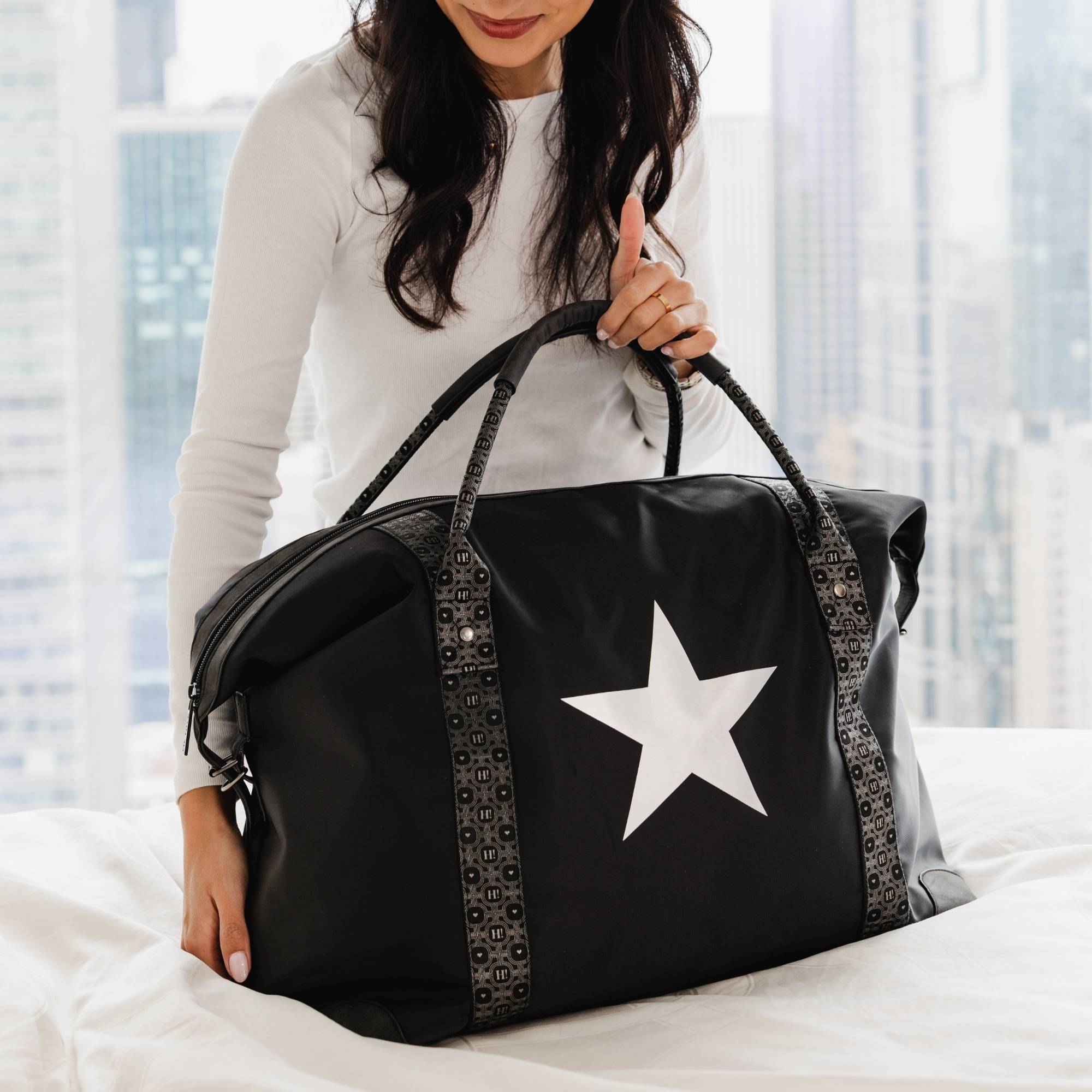 Black weekender duffle bag with silver star and signature straps styled on a bed.