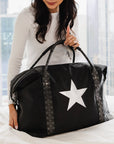 Black weekender duffle bag with silver star and signature straps styled on a bed.
