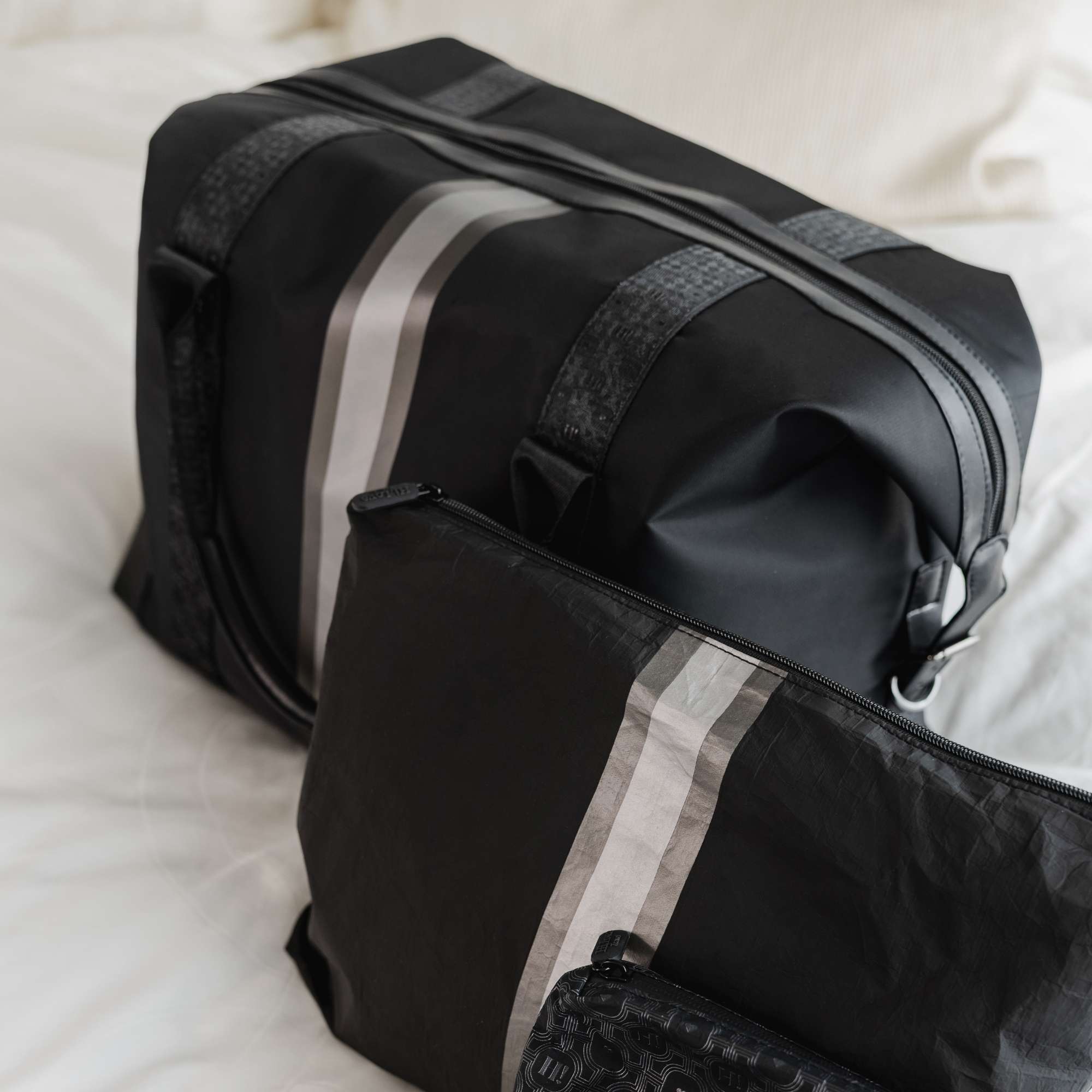 Black weekender travel bag featuring a pewter and silver center stripe, paired with a matching jumbo zipper pouch both lying on a bed.