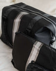 Black weekender travel bag featuring a pewter and silver center stripe, paired with a matching jumbo zipper pouch both lying on a bed.