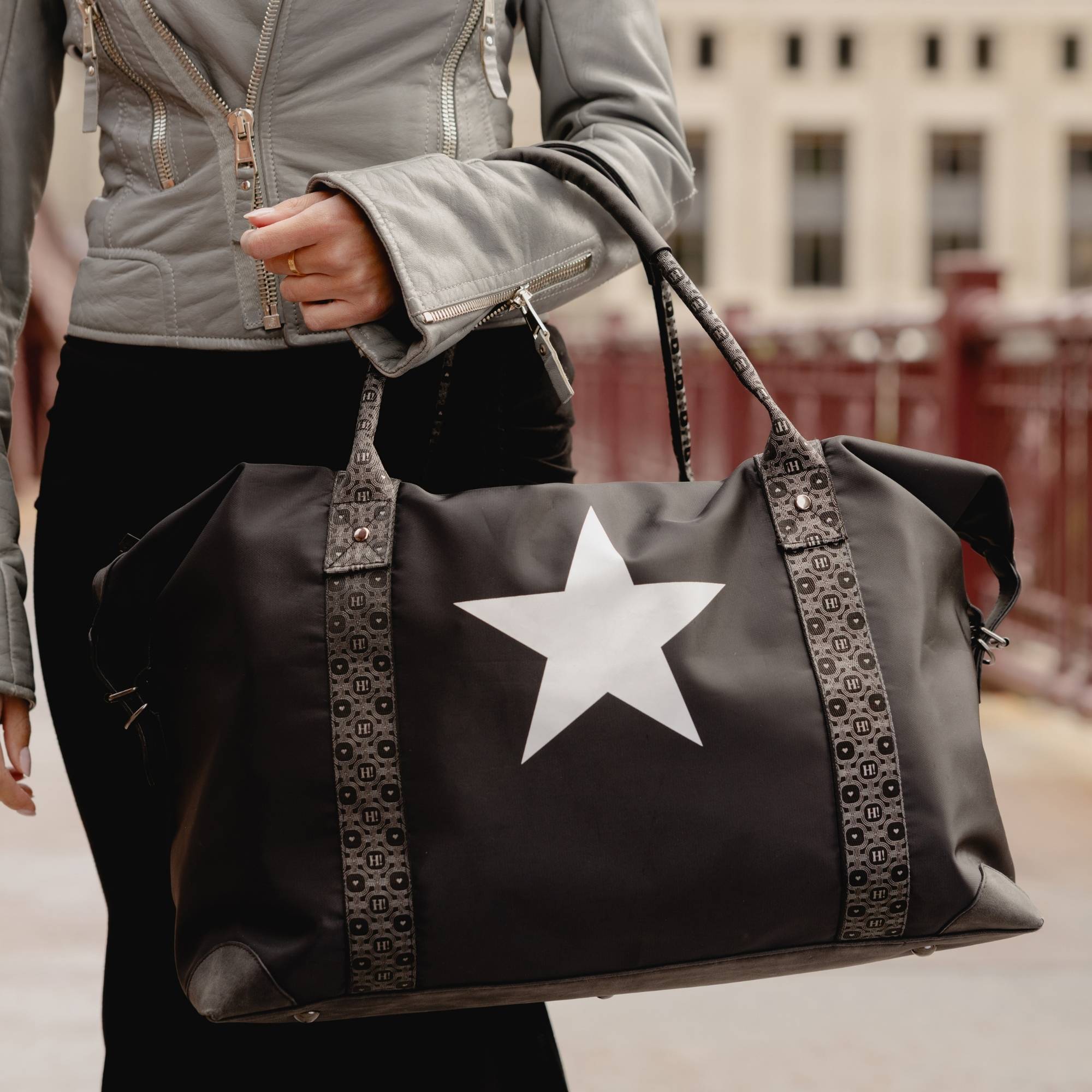 Black weekender duffle bag with silver star and signature straps carried by hand in an outdoor city setting.
