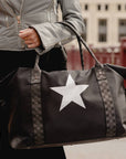 Black weekender duffle bag with silver star and signature straps carried by hand in an outdoor city setting.