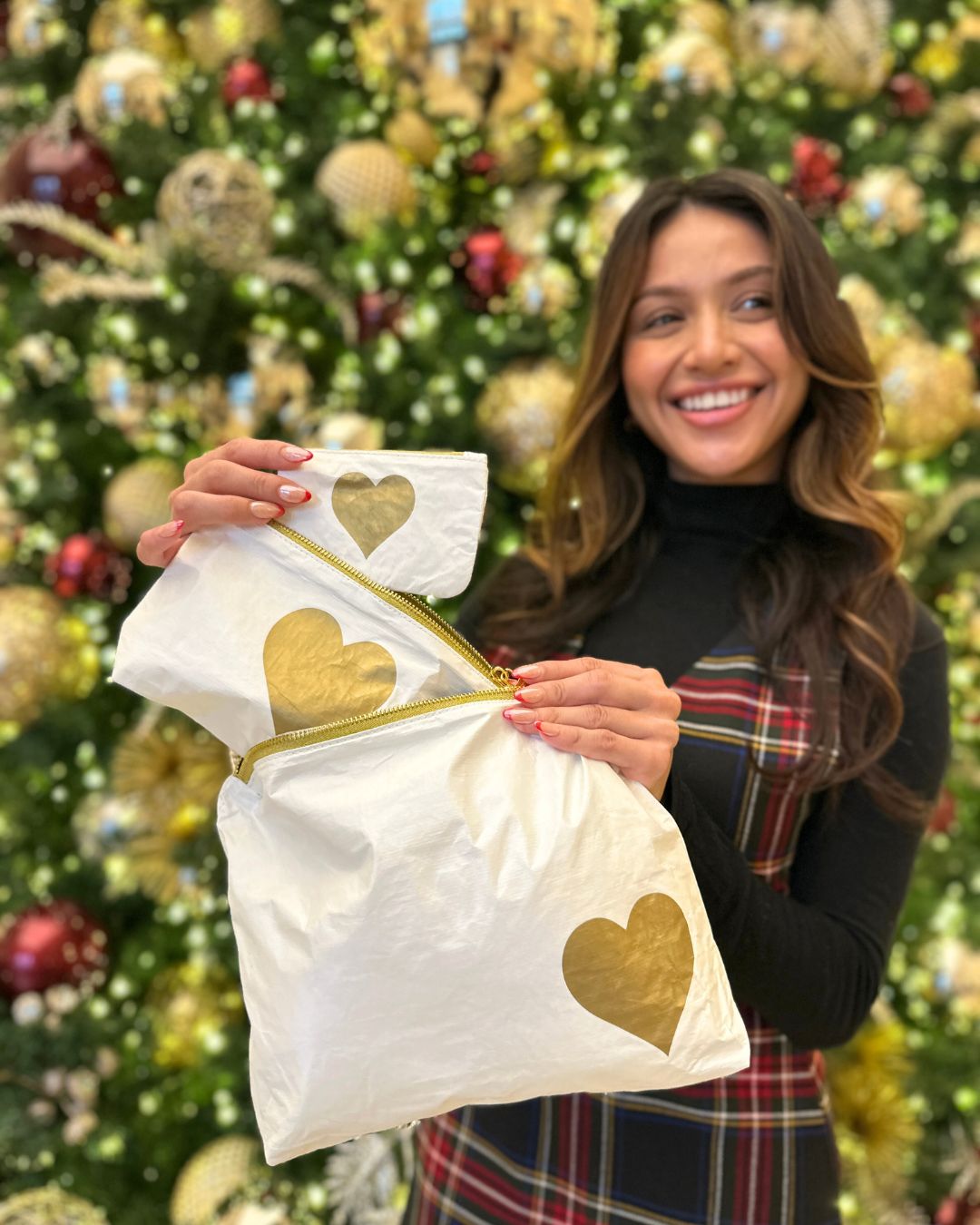 Woman holding a white zipper pouch set with gold hearts in front of a decorated Christmas tree.