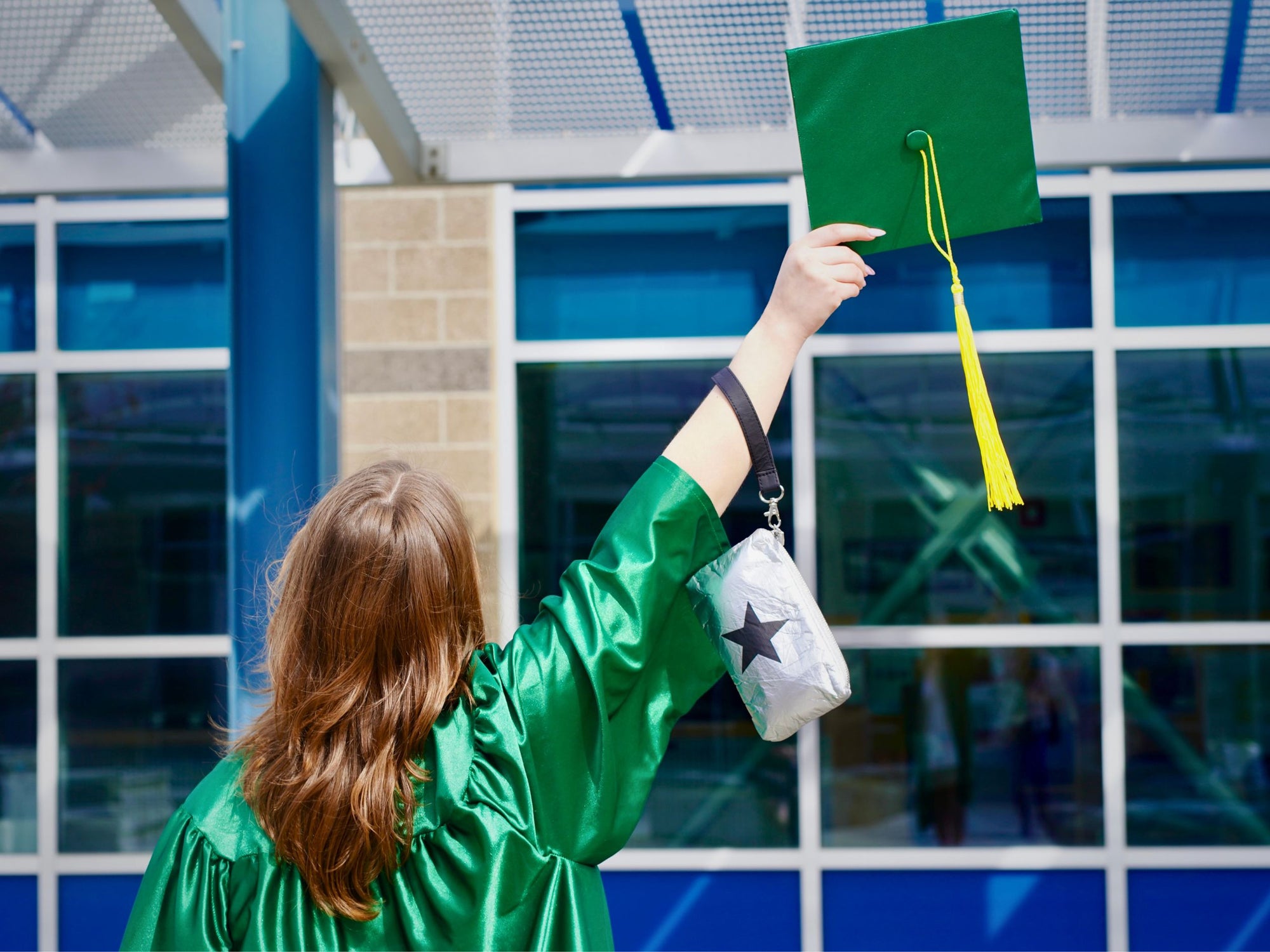 Graduate in green gown holding a green graduation cap with a silver with black start wristlet bag.