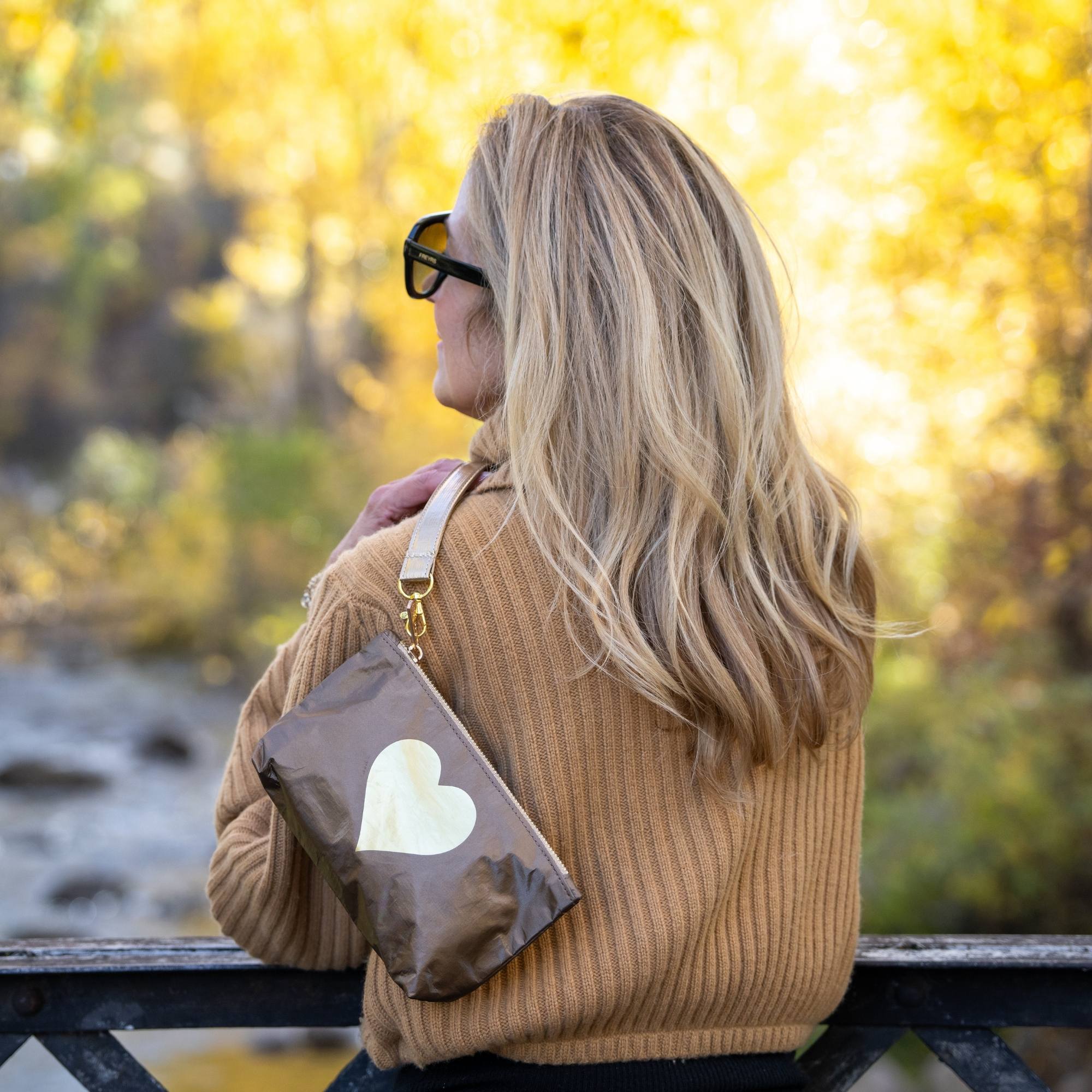 Woman wearing a bronze metallic wristlet pouch with a gold heart design over her shoulder while overlooking a river in a fall landscape.