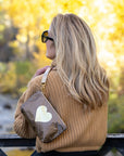 Woman wearing a bronze metallic wristlet pouch with a gold heart design over her shoulder while overlooking a river in a fall landscape.
