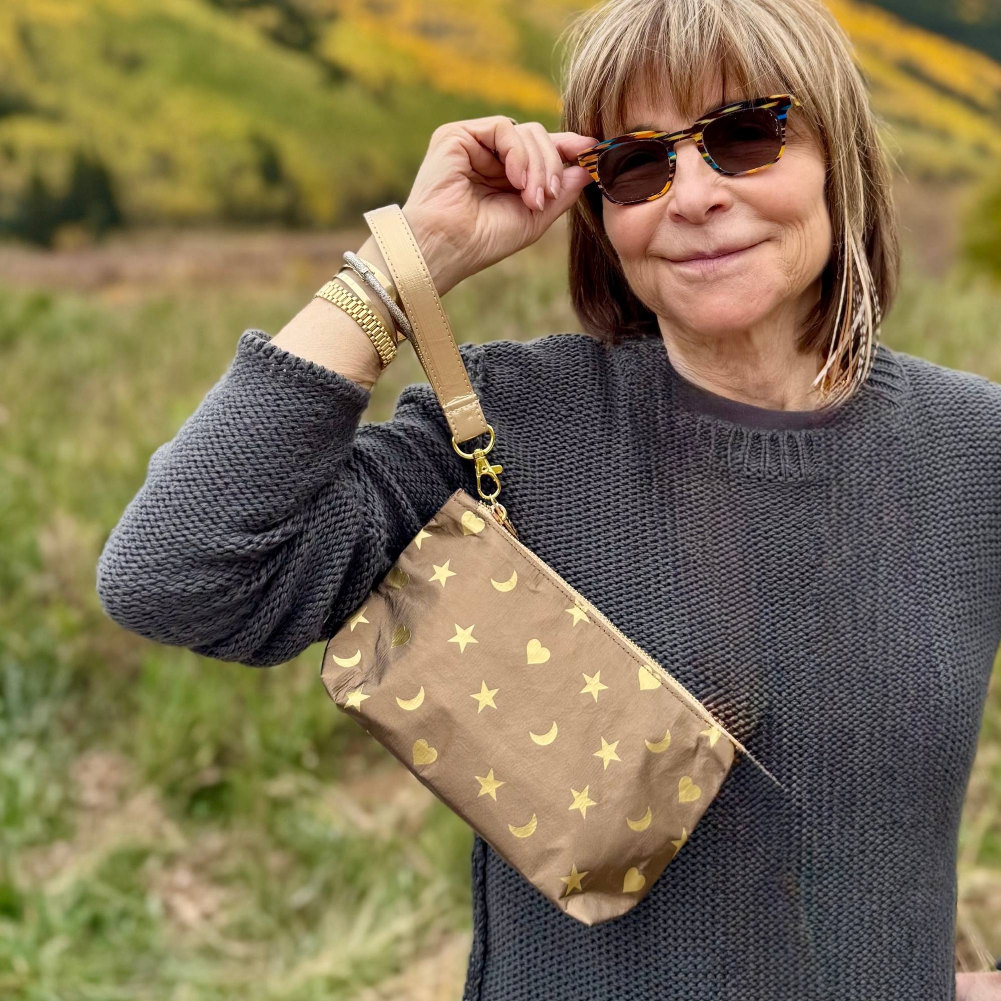 Woman wearing a bronze metallic wristlet pouch with gold heart, moon, star design outside in the fall.