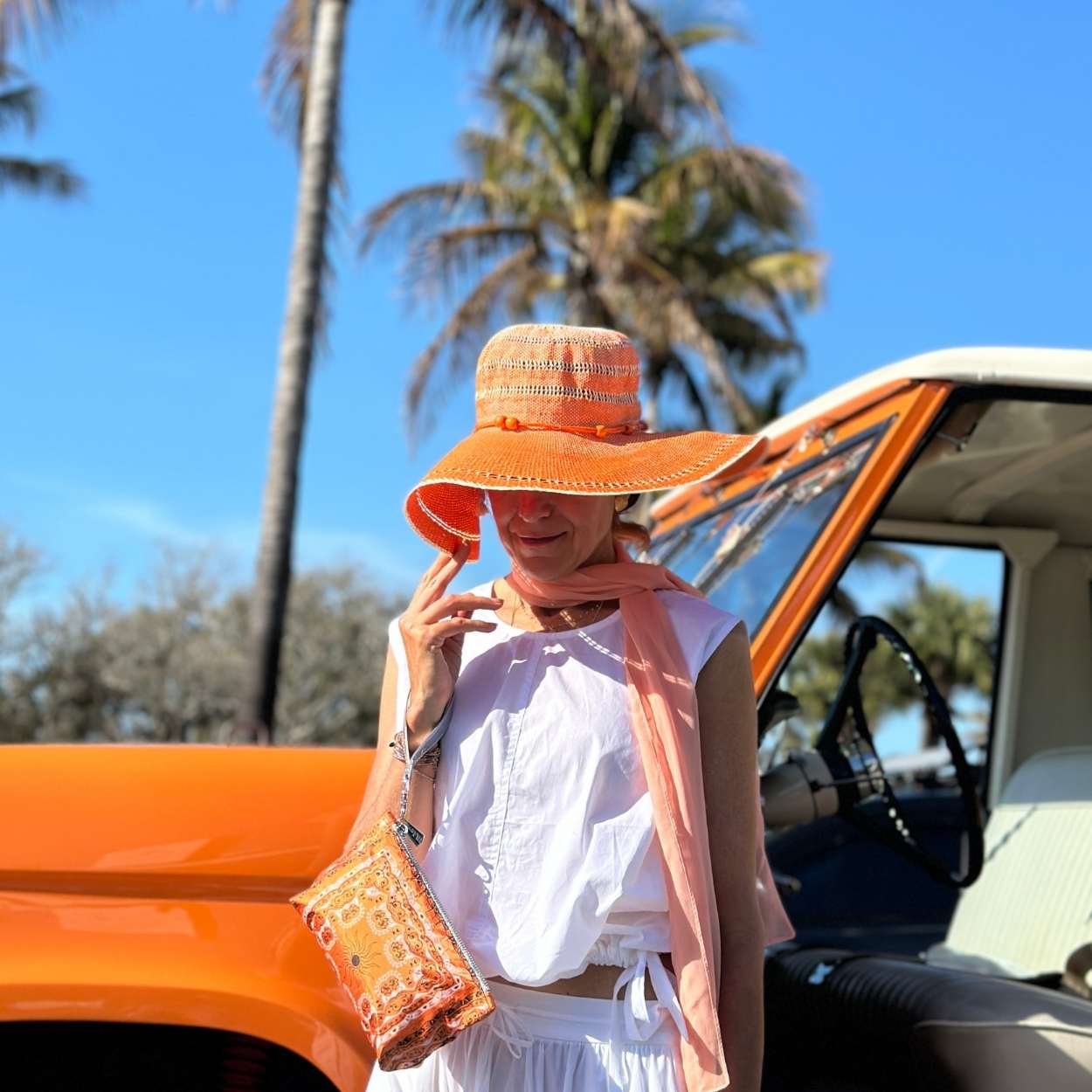 Woman wearing an orange hat, orange wristlet and white outfit standing next to a vintage car with palm trees in the background.