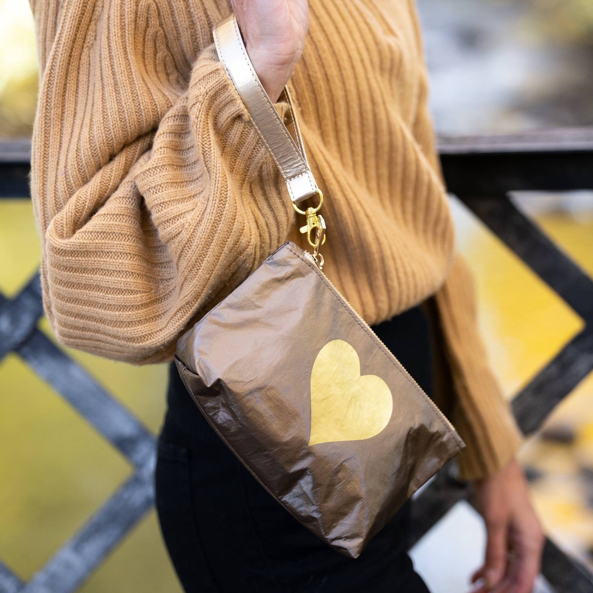 Woman wearing a bronze shimmer wristlet pouch with a gold heart design over her shoulder while overlooking a river in a fall landscape.