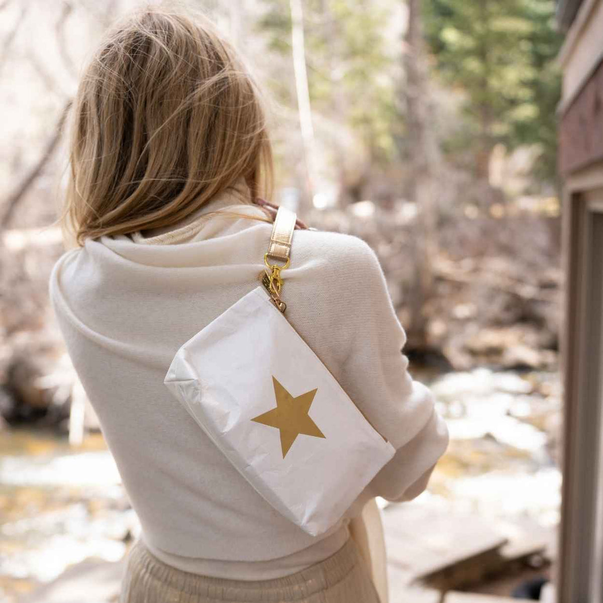 A close-up of a person wearing a cream knit sweater and holding a small white wristlet pouch with a metallic gold star on the front and a gold-toned strap in outdoor background.