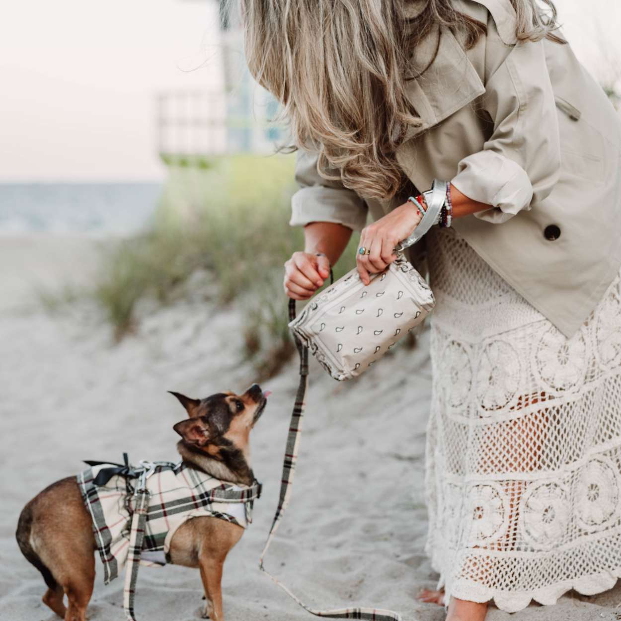 Woman in a trench coat and lace skirt holding a bandana wristlet bag, standing on a beach with a small dog in a plaid outfit.