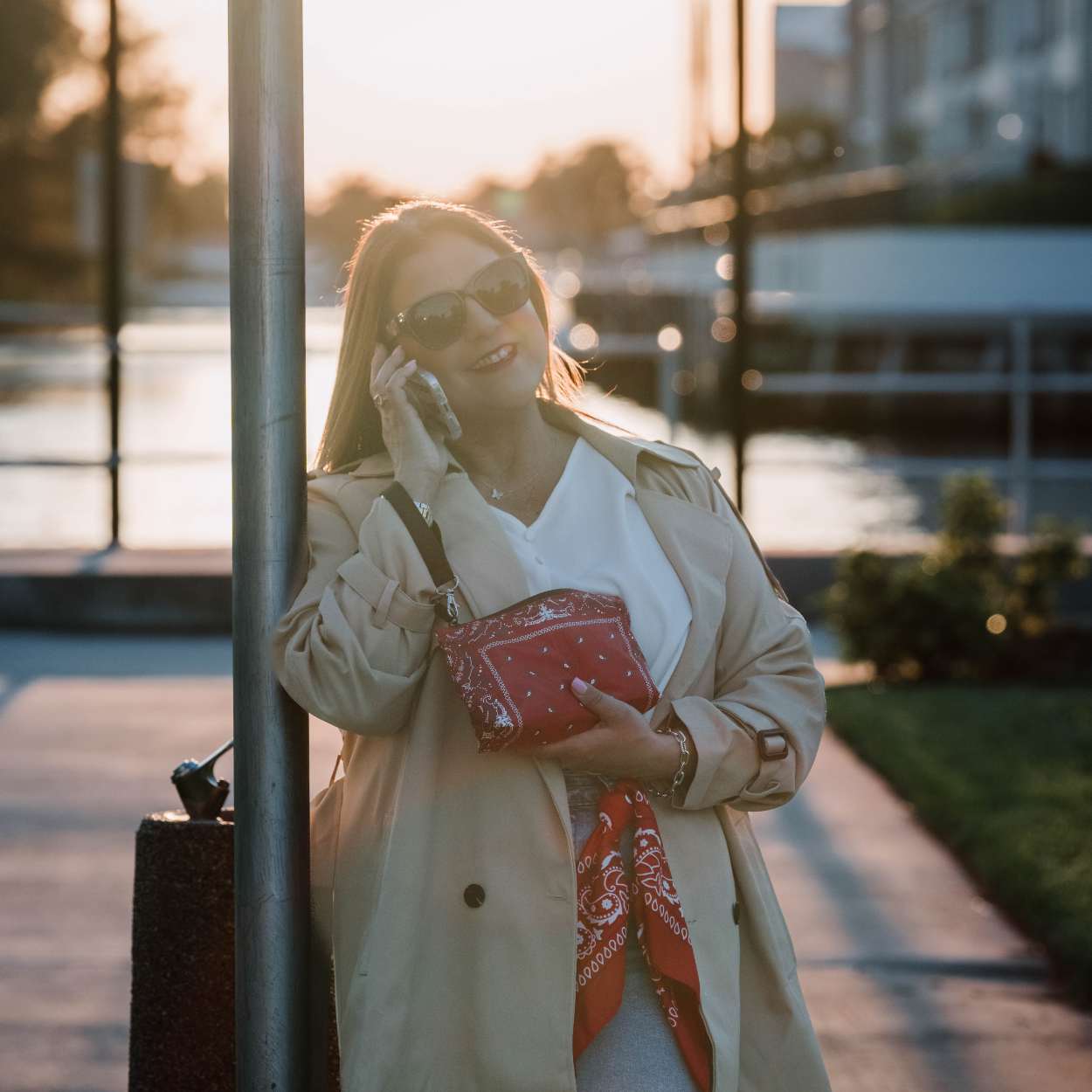 Woman in a beige coat and sunglasses holding a red bandana wristlet bag, standing next to a pole with a blurred background.