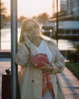 Woman in a beige coat and sunglasses holding a red bandana wristlet bag, standing next to a pole with a blurred background.
