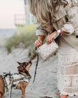 Woman in a trench coat and lace skirt holding a bandana wristlet bag, standing on a beach with a small dog in a plaid outfit.