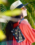 Woman wearing a straw hat and sunglasses, with a butterfly-patterned bag, against a tropical leaf background.