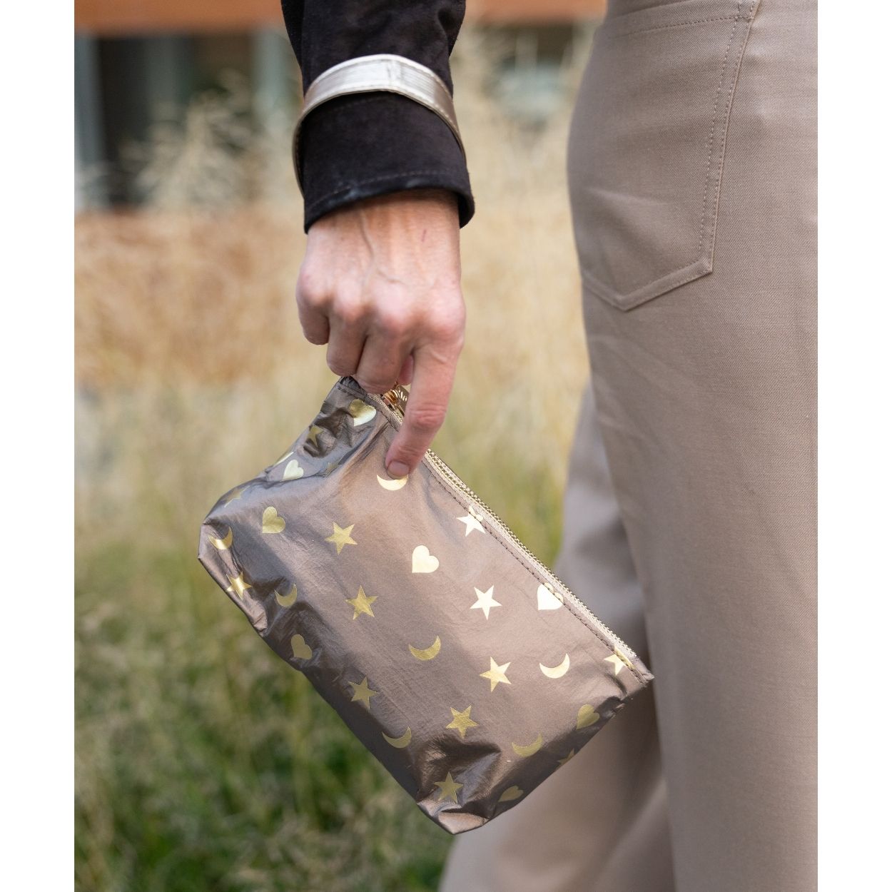 A person outdoors holding a metallic bronze wristlet decorated with gold stars, moons, and hearts. The person is wearing beige trousers and a black sleeve.