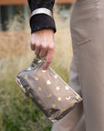 A person outdoors holding a metallic bronze wristlet decorated with gold stars, moons, and hearts. The person is wearing beige trousers and a black sleeve.