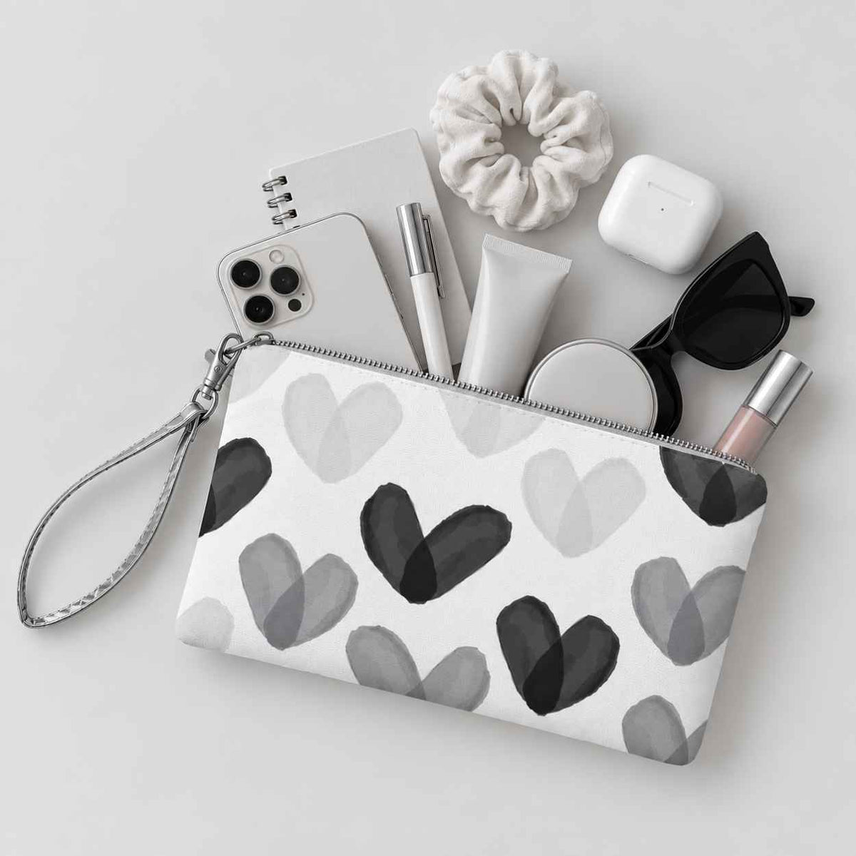 Wristlet with black and white heart print holding phone, notepad, earbuds, lip gloss, and various essentials on light background.