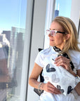 A woman with blonde hair and glasses stands by a large window, holding a white pouch decorated with a gray and black heart pattern. She is wearing a white t-shirt and a black-and-white patterned scarf, with a city skyline visible outside the glass.