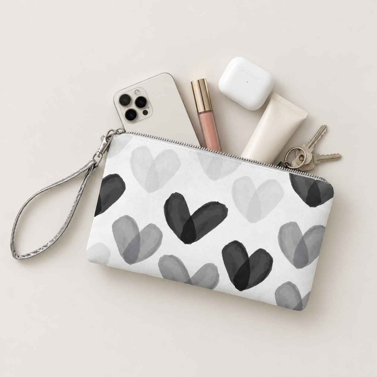 Wristlet with black and white heart print holding phone, keys, earbuds, lip gloss, and hand cream on beige background.
