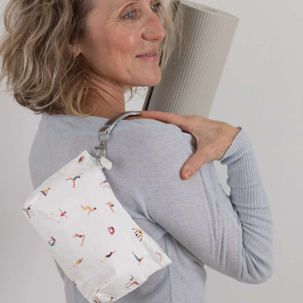 Woman holding a yoga mat with a white pouch featuring people doing yoga on her shoulder.