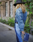 Person wearing a denim jacket and jeans holding a blue bandana wristlet with a building and plants in the background